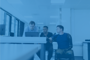 Na imagem aparecem 3 colaboradores em uma mesa de trabalho, olhando para o computador. Um deles é uma pessoa com deficiência, usuário de cadeira de rodas, reforçando o tema central do post.