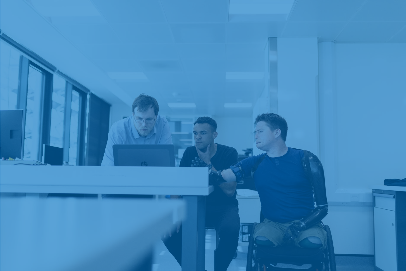 Na imagem aparecem 3 colaboradores em uma mesa de trabalho, olhando para o computador. Um deles é uma pessoa com deficiência, usuário de cadeira de rodas, reforçando o tema central do post.
