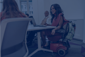 Na imagem, temos uma mulher em uma cadeira de rodas motorizada. Ela está em uma mesa de um escritório, próxima de outros 2 colegas de trabalho, representando a diversidade.