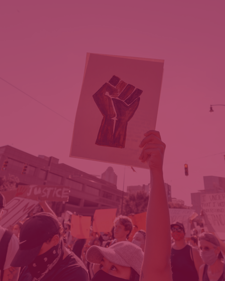 A imagem apresenta uma pessoa levantando uma placa, no meio de uma multidão, no que aparenta ser um protesto. Na placa, desenhado, há um punho cerrado, símbolo do movimento de luta contra o racismo.