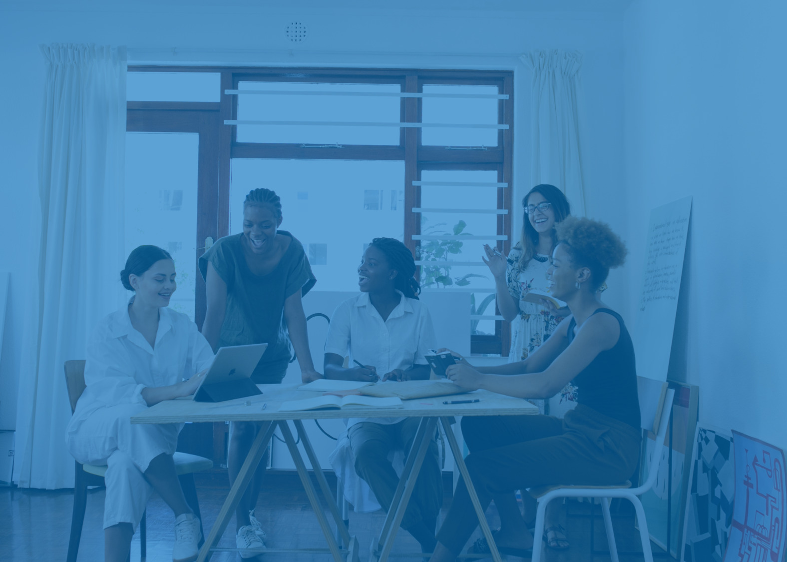 A imagem mostra um grupos de trabalho, em volta de uma mesa, formado por mulheres sorrindo.