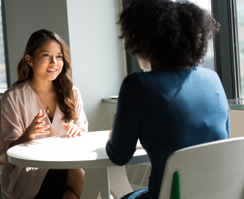 Na imagem, duas mulheres conversando ao redor de uma mesa branca.