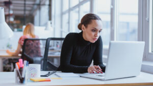 Na imagem, uma mulher negra trabalhando olhando para o computador de forma concentrada.