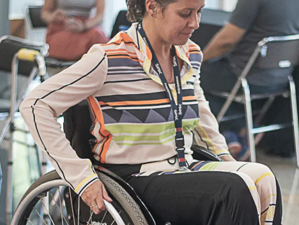 Na imagem, destaque em primeiro plano uma mulher usando cadeira de rodas para se locomover em um ambiente.