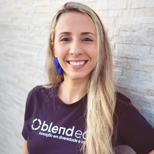 Marcela é uma mulher loira, de cabelos longos. Está vestindo uma blusa preta com o logo da Blend Edu. Ao fundo está uma parede de pedras brancas.