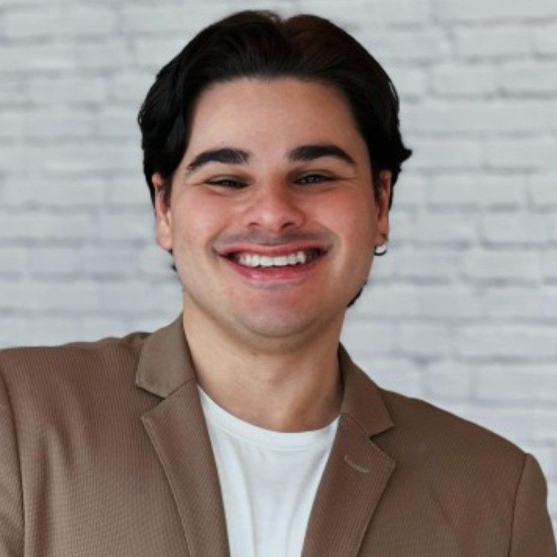 A imagem conta com uma pessoa não binária branca, usando um pequeno brinco de argola na orelha esquerda. A pessoa veste um blazer em um tom de marrom média e uma blusa branca por baixo. Atrás tem uma parede branca com tijolos.