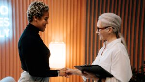Duas mulheres apertando as mãos após um processo de recrutamento e seleção. Uma jovem e negra. Uma sênior e branca.