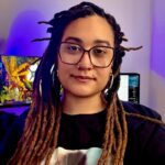 Mulher negra de pele clara, usa óculos, com dreadlocks longos e piercing no nariz, olha para a câmera com expressão tranquila. Ao fundo, monitores de computador e iluminação em tons de roxo e azul.