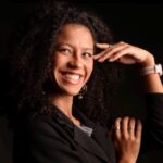 Mulher, parda de cabelos cacheados soltos, sorrindo para a câmera. Está de lado, com uma das mãos apoiada próxima ao rosto. Veste blazer preto, com anéis e relógio claro no pulso. Fundo preto com iluminação que destaca seu rosto e expressão sorridente.